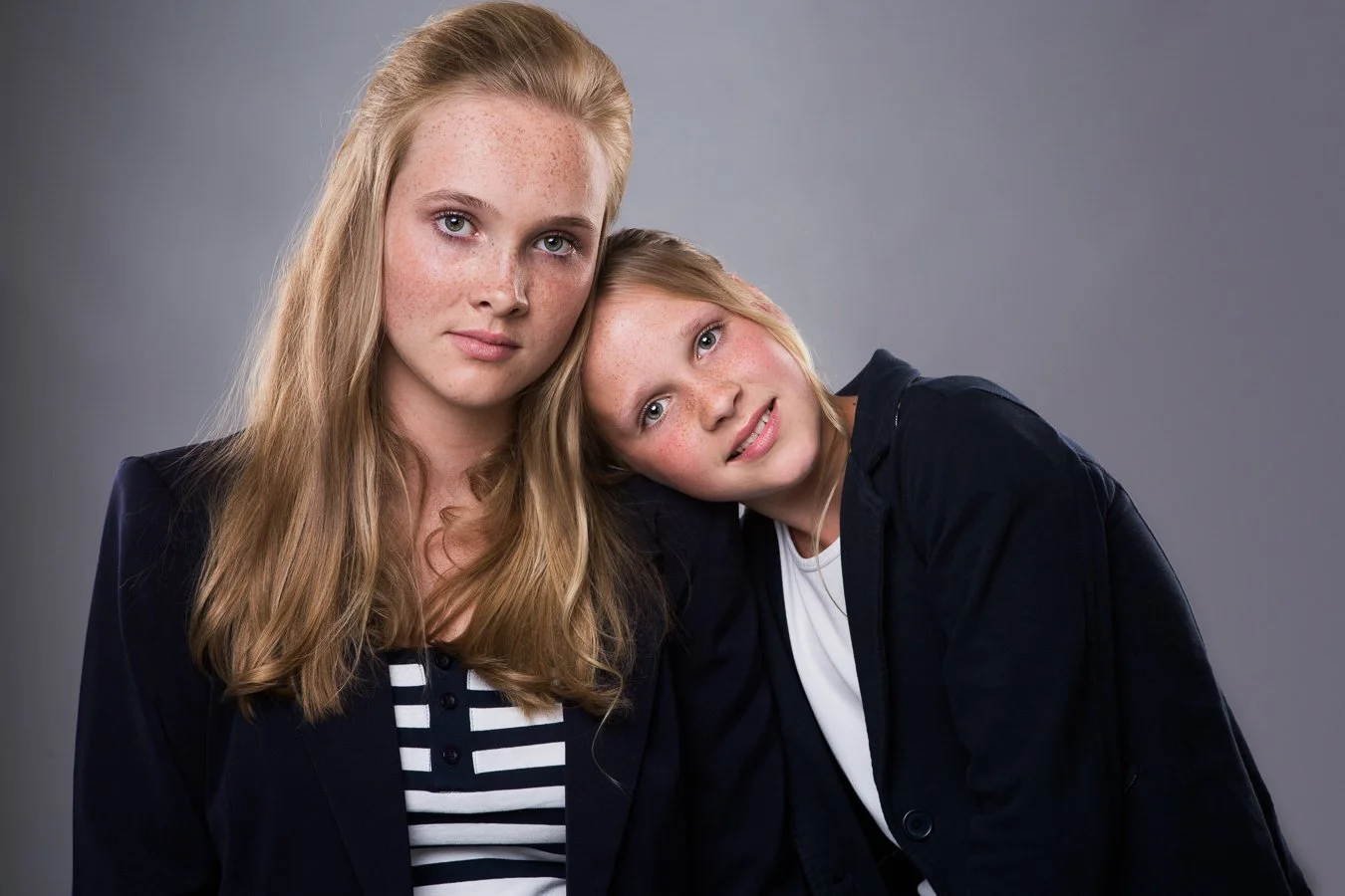 Twee jonge meisjes met lichtbruine en blonde haar, gekleed in donkerblauwe jassen en witte shirts, poseren tegen een grijze achtergrond.