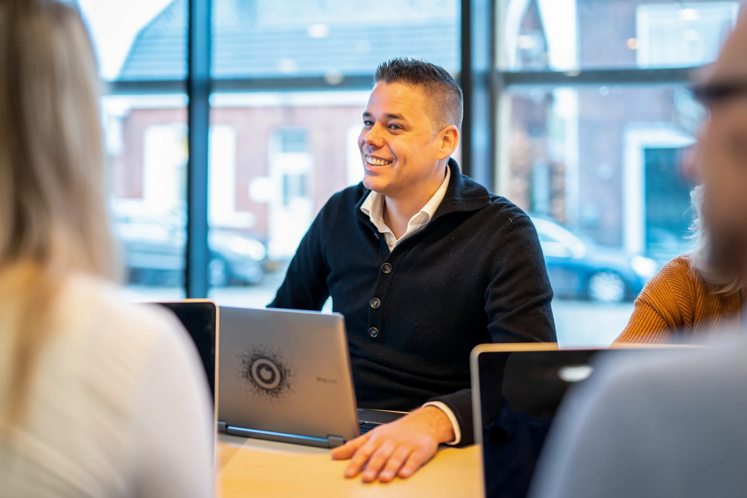 Een man met korte haar, zittend aan een tafel in een vergaderruimte, lachend en in gesprek met andere mensen, omgeven door laptops.