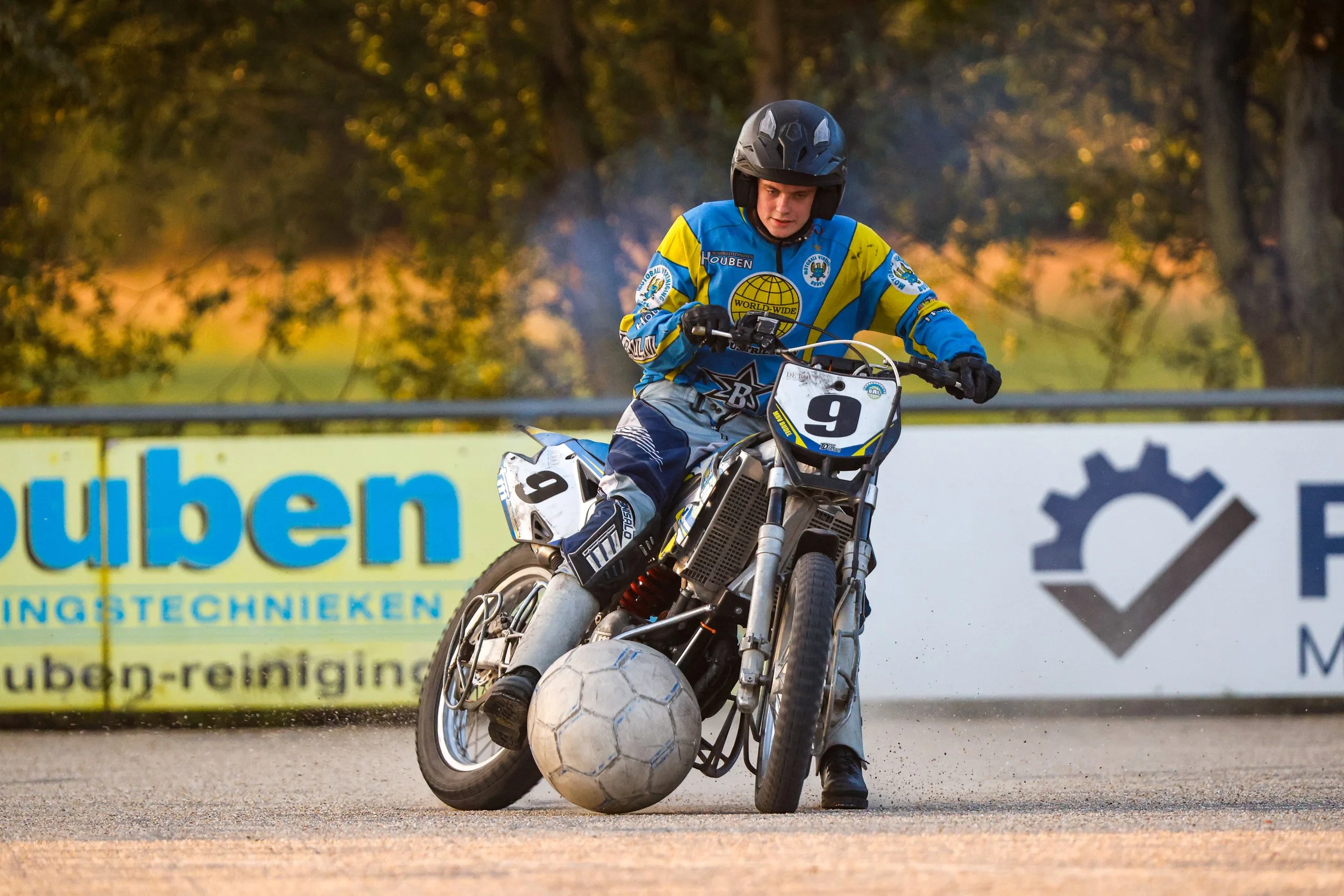 Een jongen in een blauwe en gele sportoutfit speelt motorsport met een aangepaste motor op een veld, terwijl hij een voetbal met een motorrijwiel raakt. Motoball in Budel