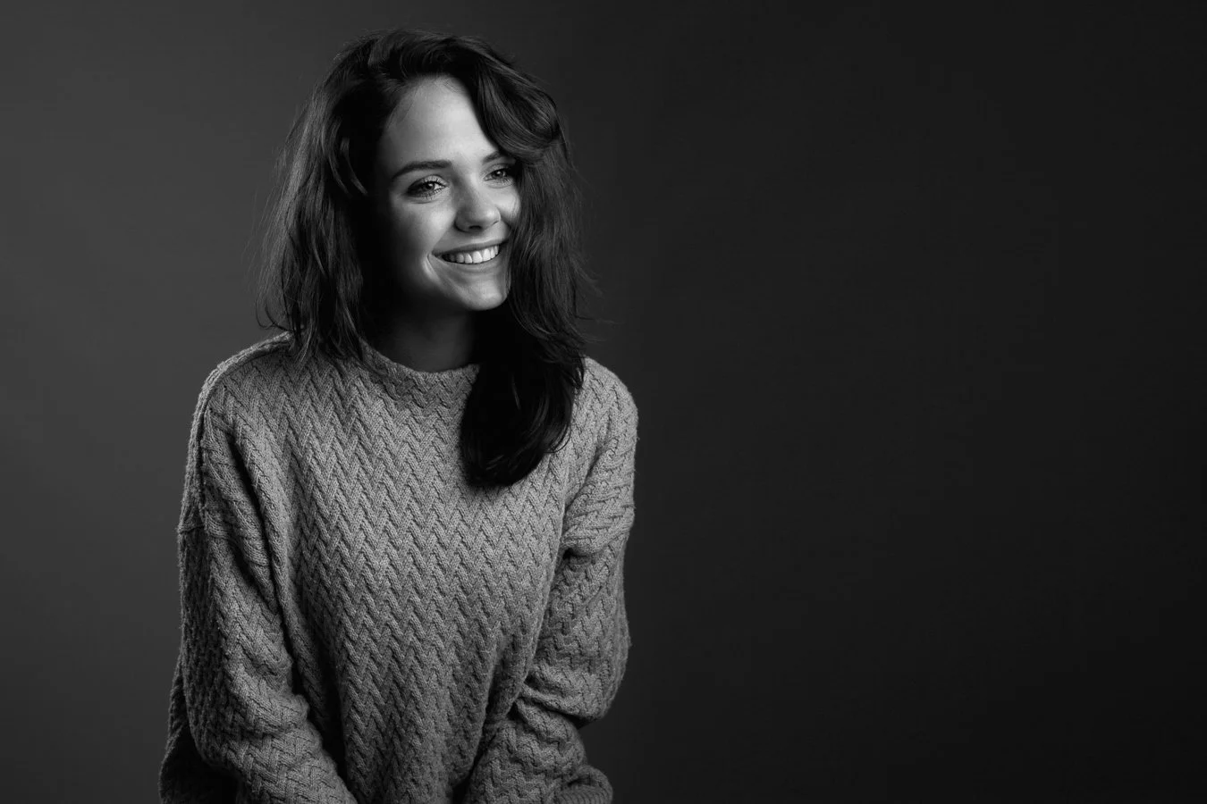 Zwart-wit foto van een lachende vrouw met steil haar, gekleed in een gebreide trui, tegen een effen achtergrond.