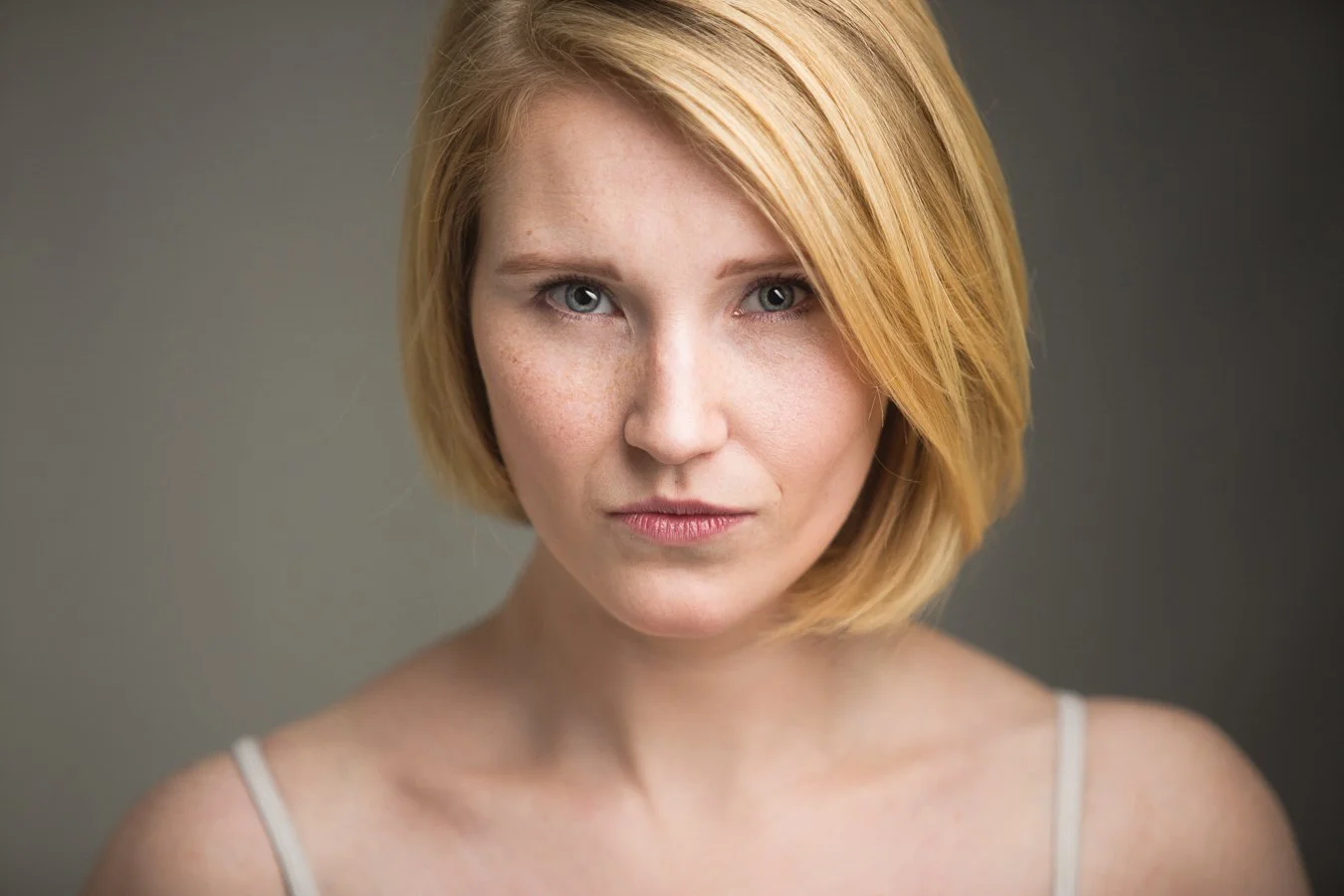 Portret van een vrouw met rood haar, blauwe ogen en een neutrale uitdrukking, gedragen in een lichte top, tegen een grijze achtergrond.