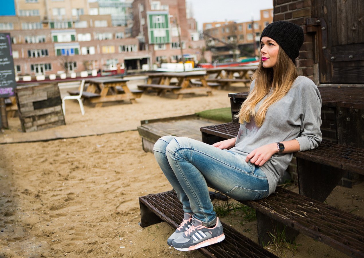 Een jonge vrouw zit op een houten bankje buiten, gekleed in een grijze hoodie, blauwe jeans en sportschoenen, met een zwarte muts en een horloge, in een stedelijke omgeving met gebouwen op de achtergrond.