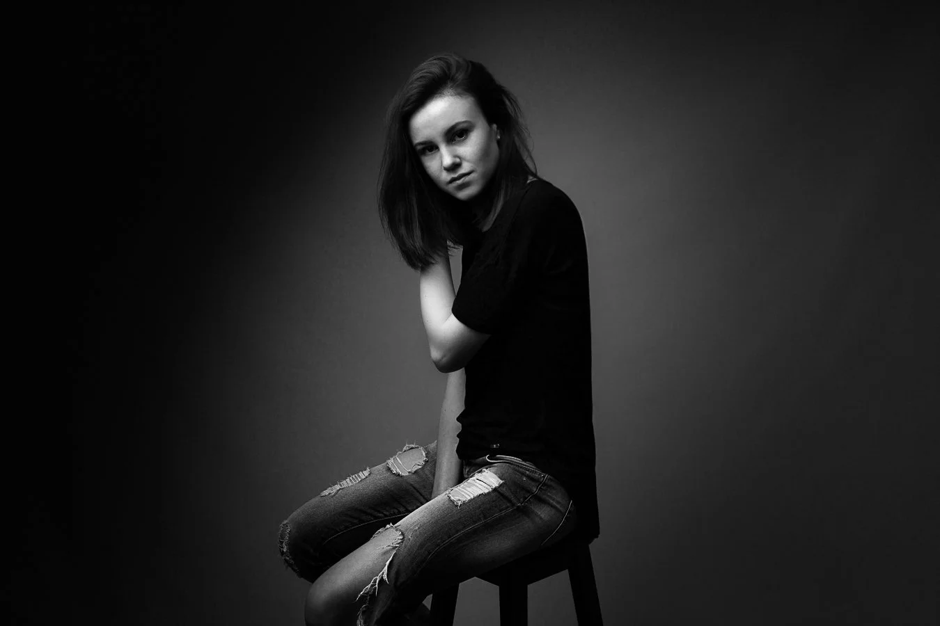 Zwarte en witte foto van een jonge vrouw met donker haar, zittend op een kruk, met een rustige uitdrukking, in een donkere kamer met egale achtergrond.