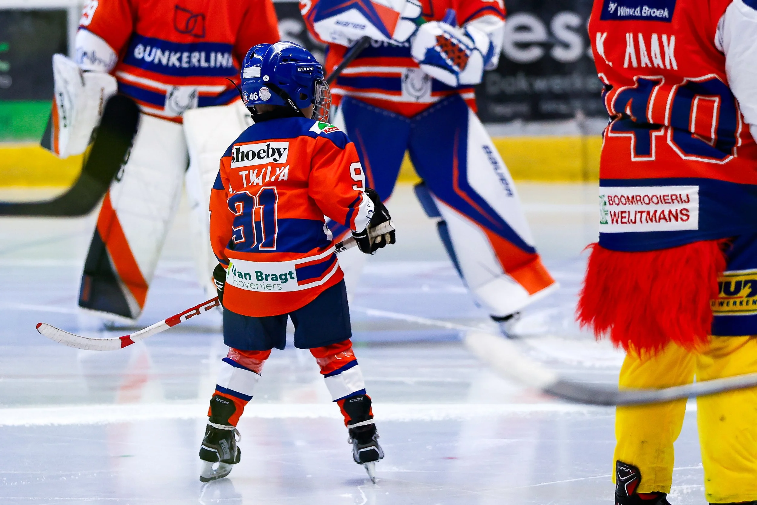 Jeugd hockey spelers tijdens een wedstrijd op het ijs, gekleed in gele, blauwe en rode uniforms, met een goalie in witte uitrusting.