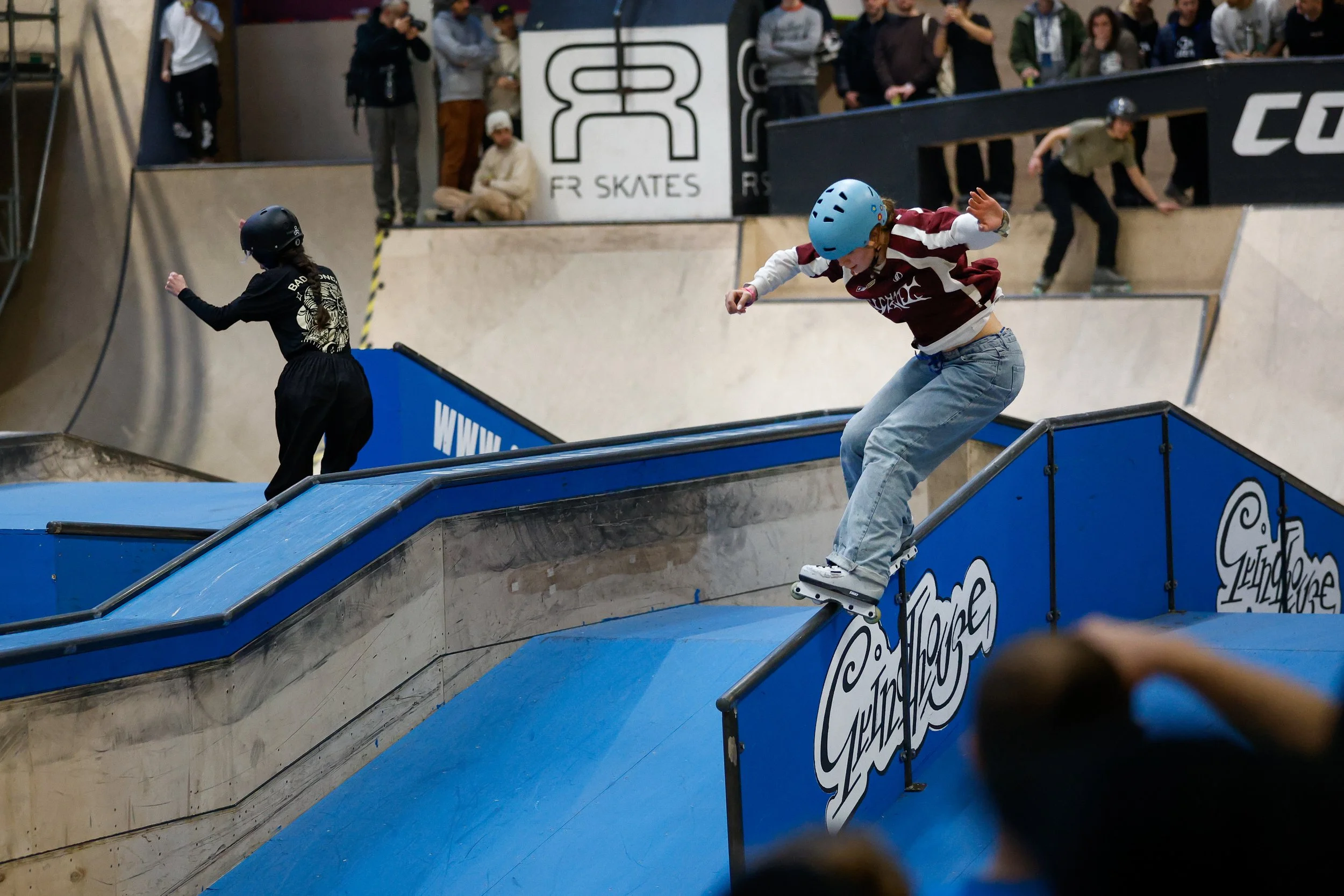 Een jonge skater op inlineskates met een blauwe helm en een maroon en wit shirt voert een trick uit op een skatebaan binnen een skatepark, terwijl andere skaters en toeschouwers toekijken.