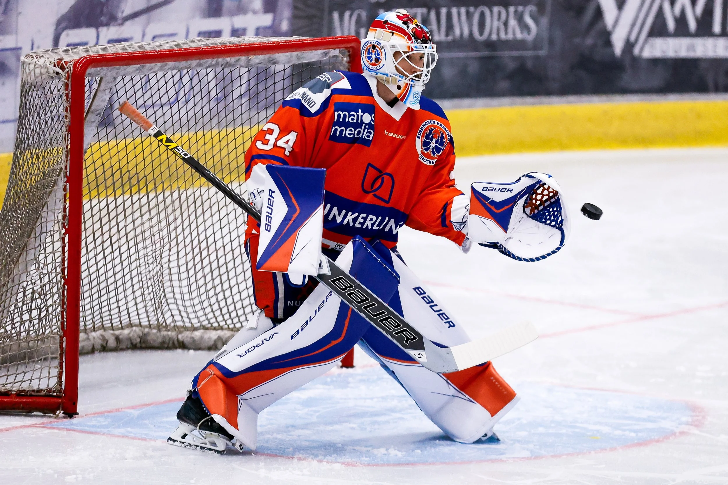 IJshockeykeeper in een oranje, blauwe en witte uniform staat voor het doel, bereidt zich voor om een schot te blokkeren. De speler draagt een masker en uitrusting met het merk Bauer en de kleuren van het team Eindhoven Kemphanen Icehockey.
