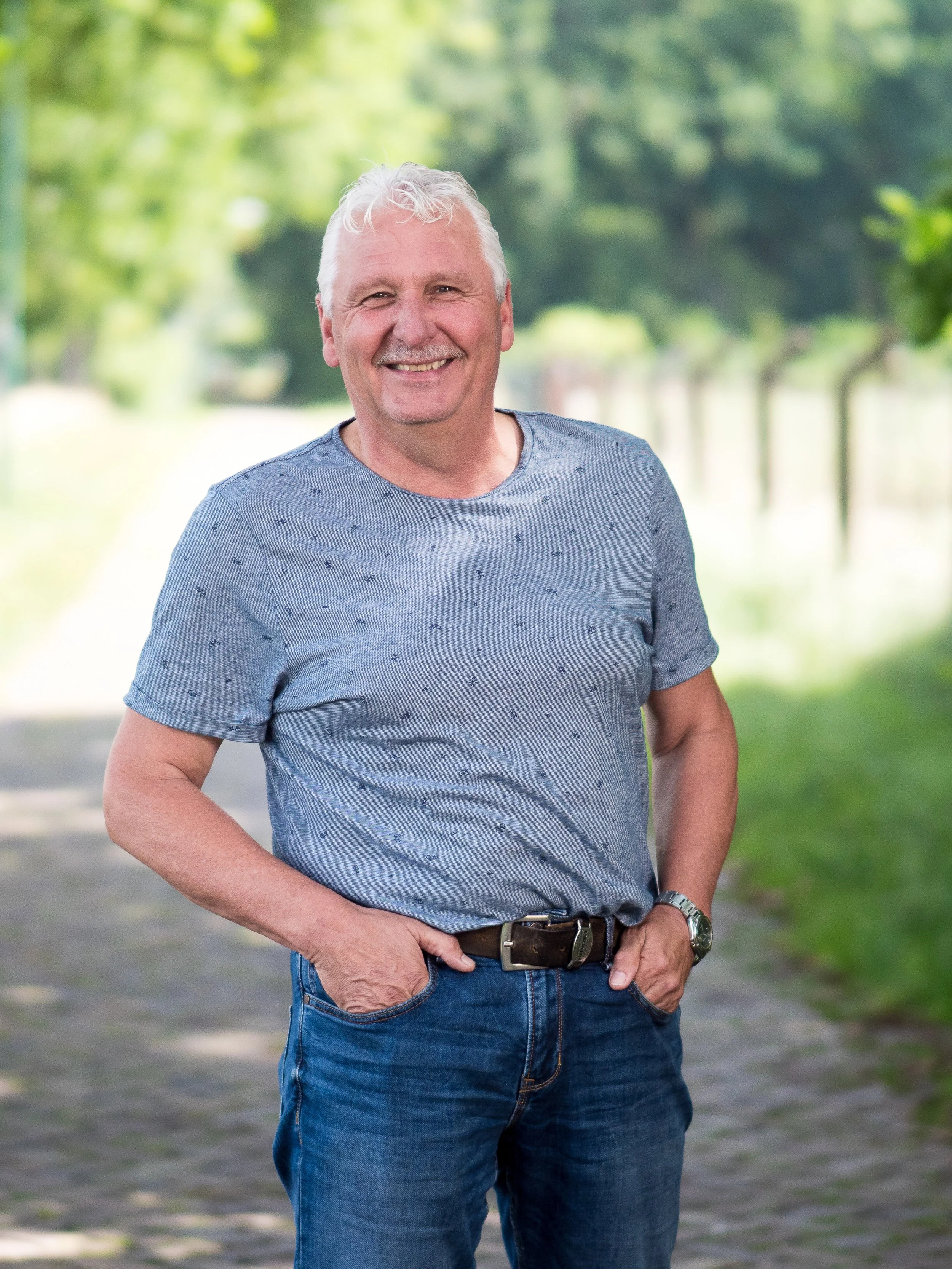 Een lachende man met grijs haar in een blauwe T-shirt en blauwe jeans, wandelend op een pad in een park met groene bomen.