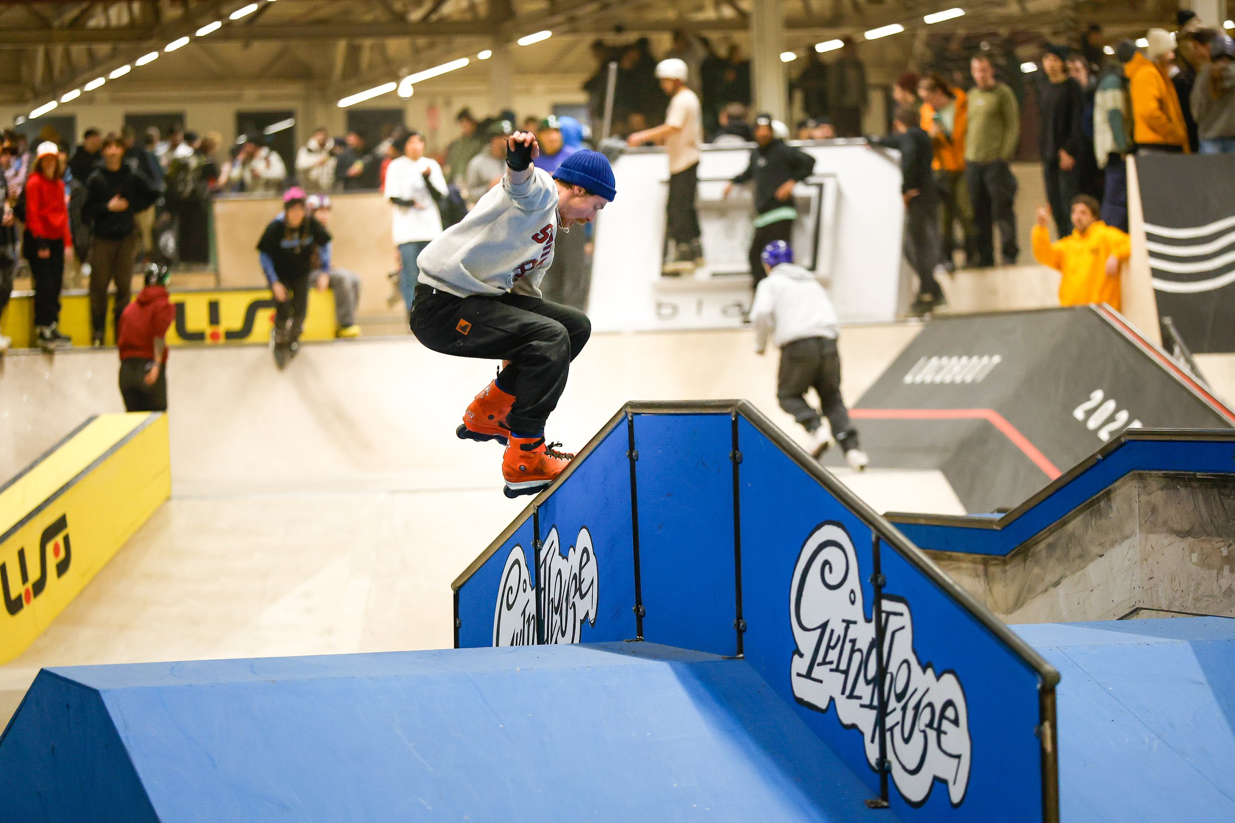 Een inlineskater voert een trick uit op een blauwe rail in een binnen skatepark Area 51 Eindhoven met een groep toeschouwers op de achtergrond. tijdens winterclash