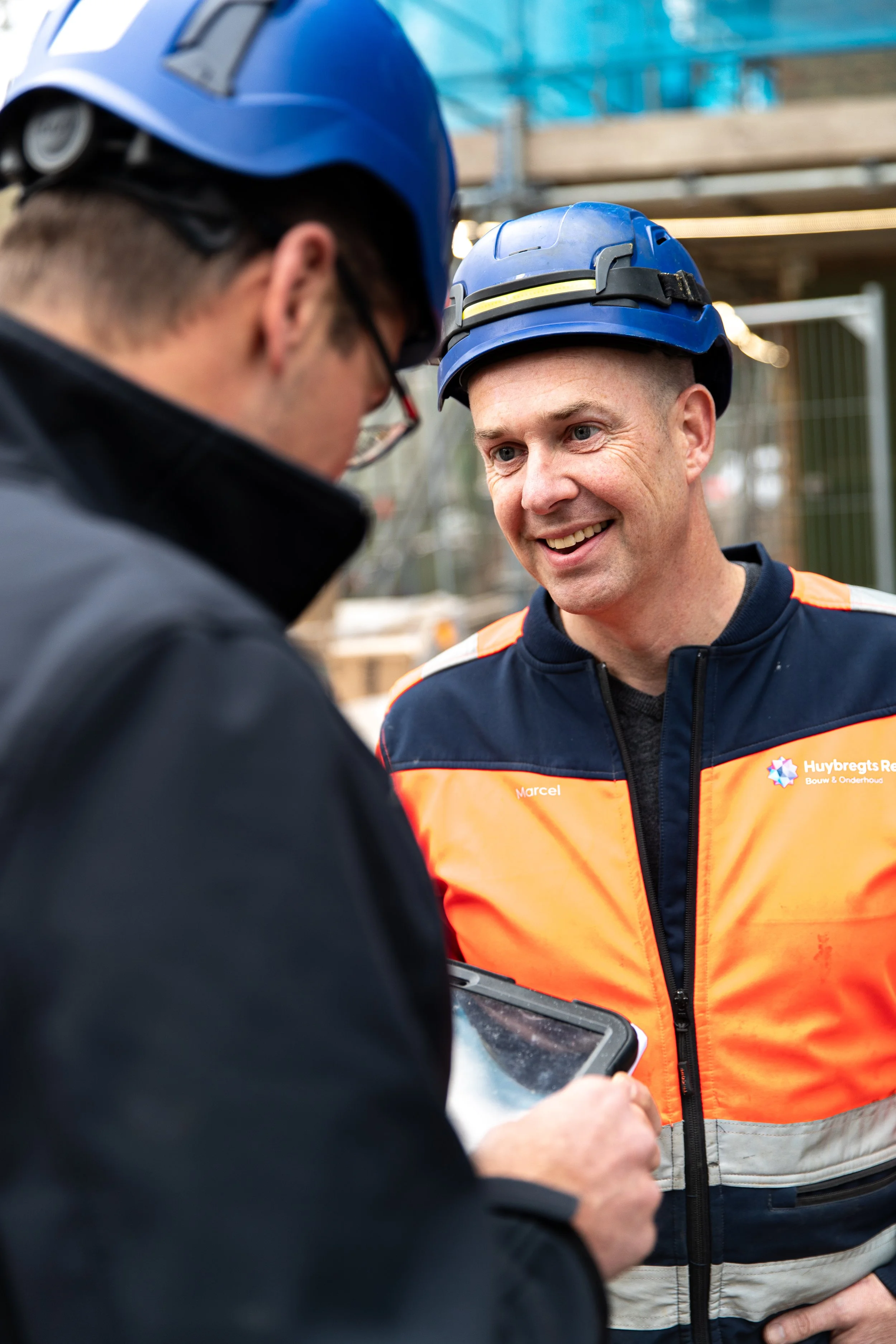 Twee bouwvakkers met helmen praten op een bouwplaats, één wijst naar een tablet, lachend.