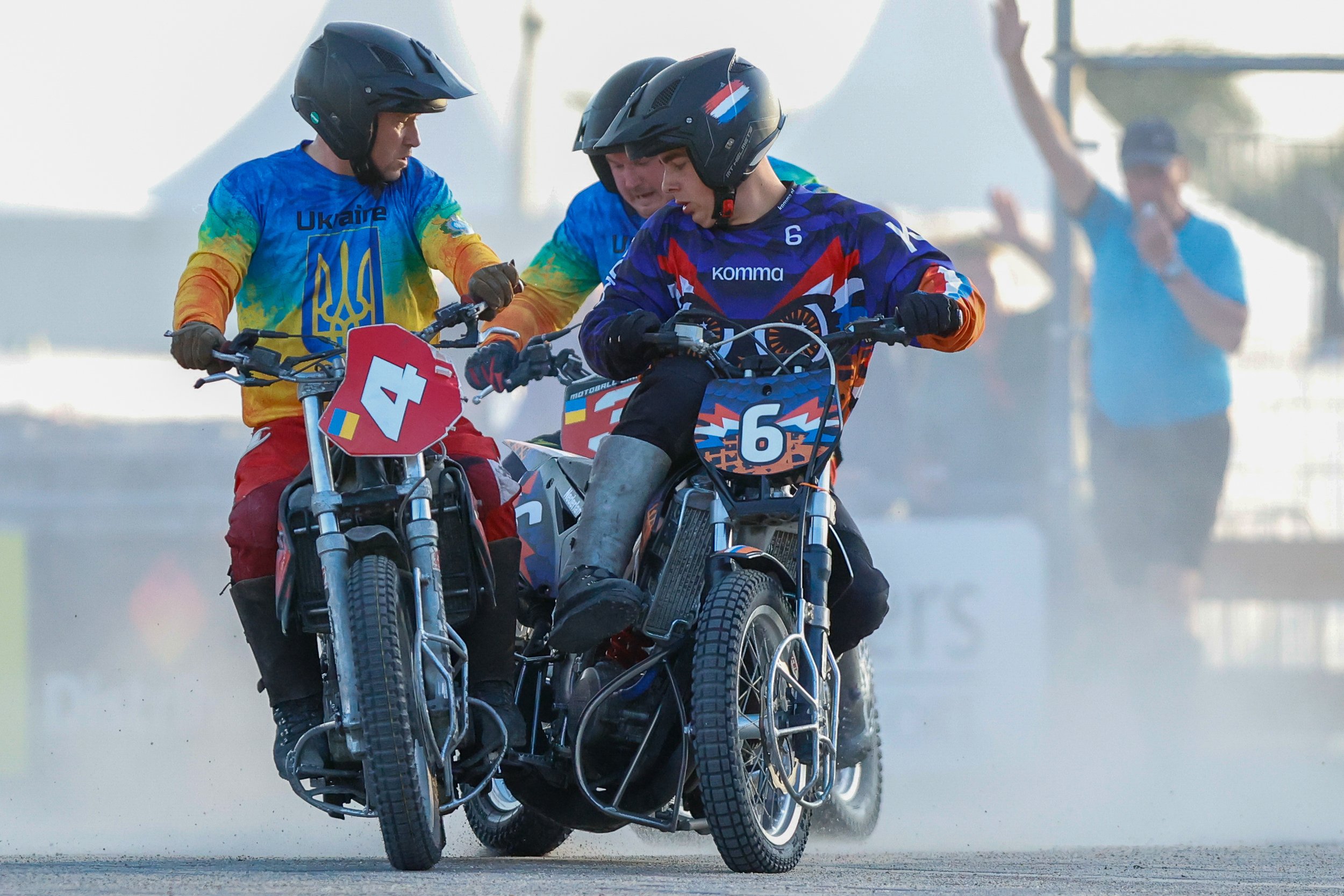 drie motoball spelers in een race, met helmen en beschermende uitrusting, op sportfietsen met nummerplaten, op een asfaltbaan met stof en rook.