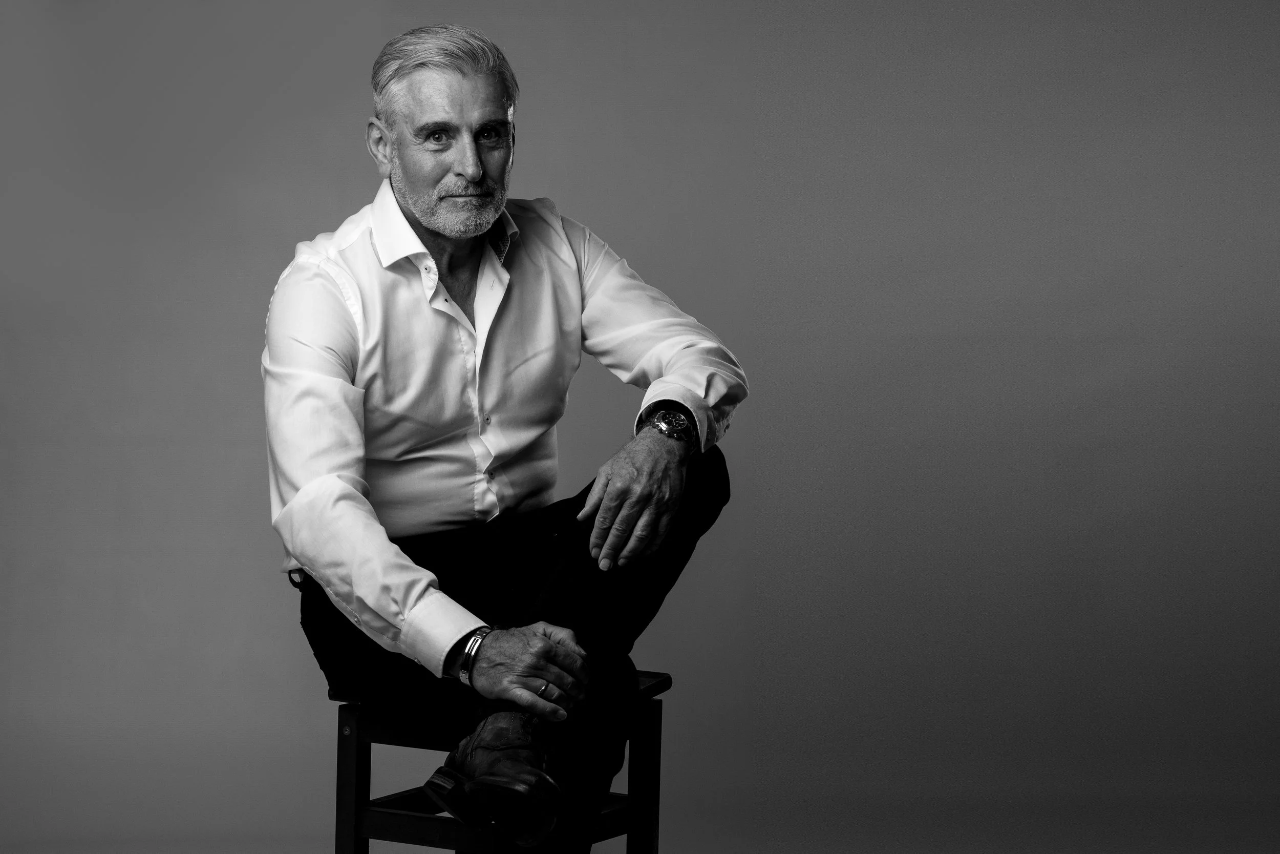 Zwarte en witte foto van een oudere man met een lange mouw witte overhemd, zittend op een stoel, met een neutrale achtergrond.