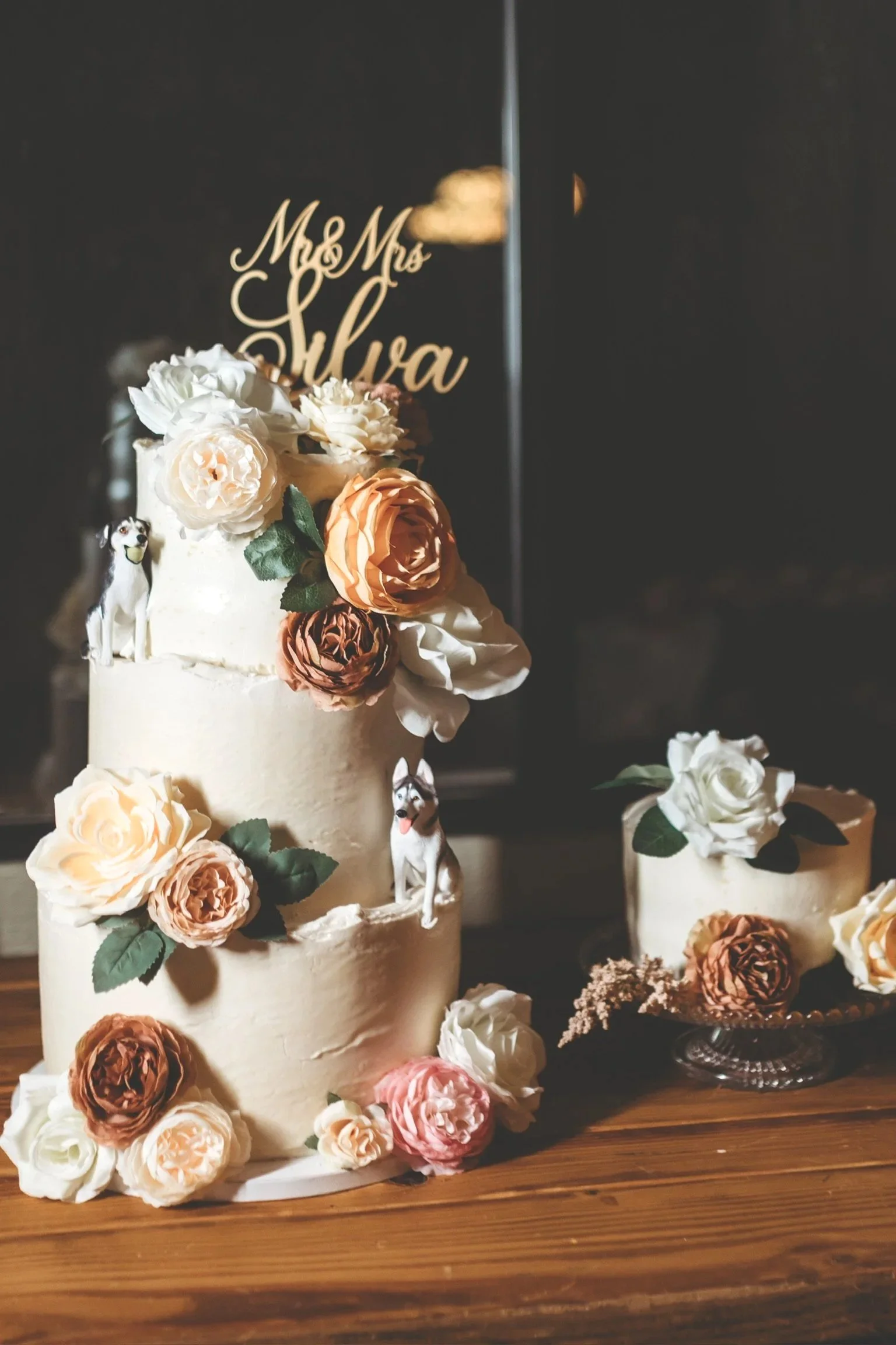 Three-tier white wedding cake decorated with various pink, white, and deep red flowers, including roses, placed on a wooden board.
