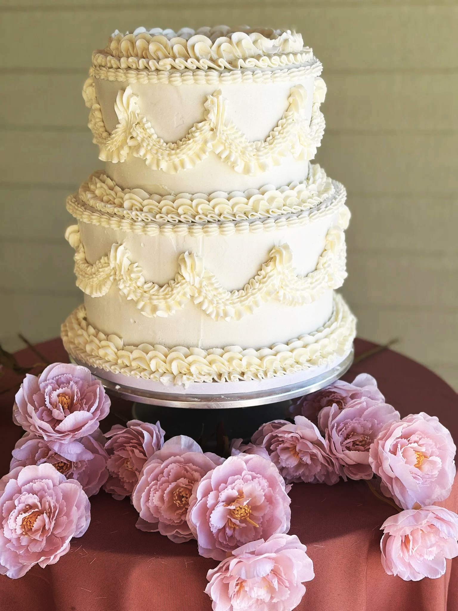 White frosted cake topped with pink chrysanthemums and green leaves, set on a silver tray with a gold cake knife and server on a white cloth napkin next to it.