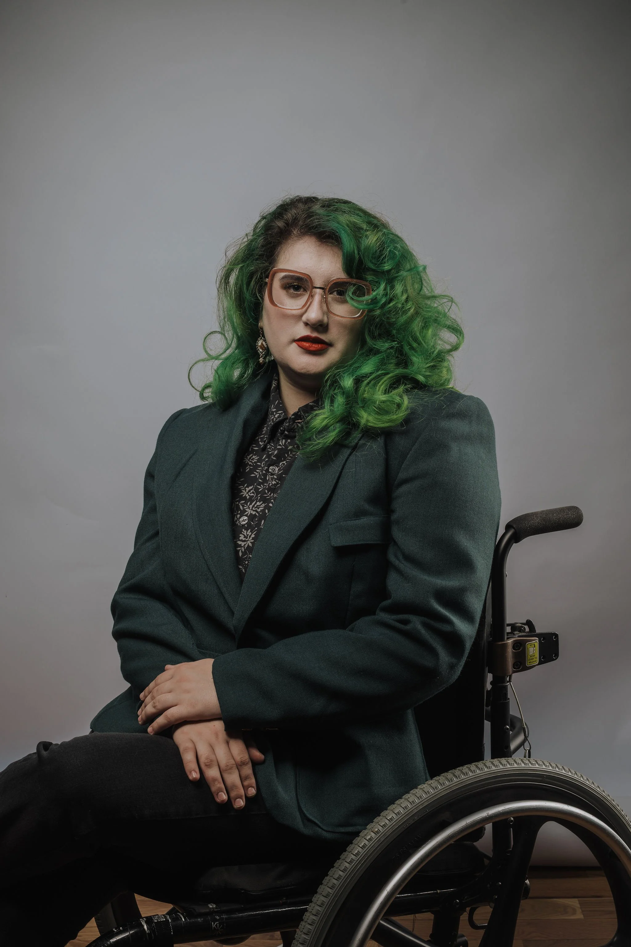 Amelia: a white woman with green hair and red lipstick, sits in her wheelchair with her arms crossed in her lap. she is modeling a forest green blazer with one breast pocket and large shoulderpads.