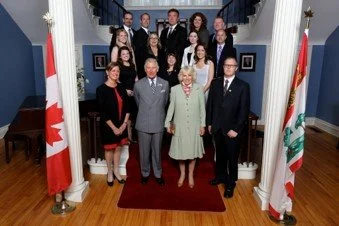 Planning committee and Their Royal Highness' in 2014 standing on a staircase  with two flags on either side, in front of a blue wall with artwork.