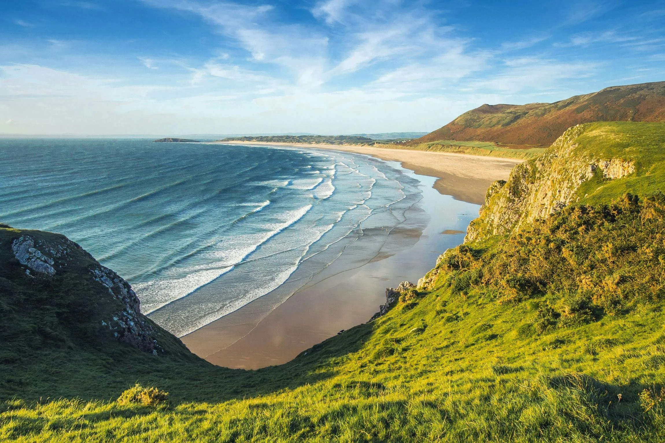A coastal landscape with grassy cliffs, a sandy beach, and gentle ocean waves under a blue sky with some clouds.