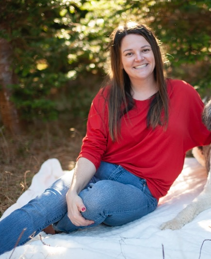 A woman with long dark hair, wearing a red shirt and blue jeans, sitting outdoors on a blanket, smiling at the camera with sunlight filtering through trees in the background.