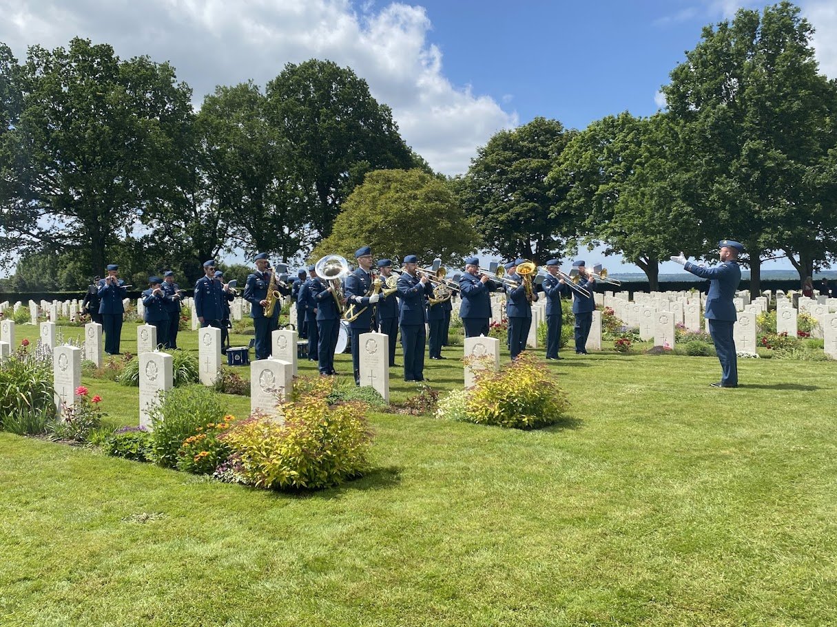 A military band performing at a cemetery with headstones, trees, and a partly cloudy sky in the background.