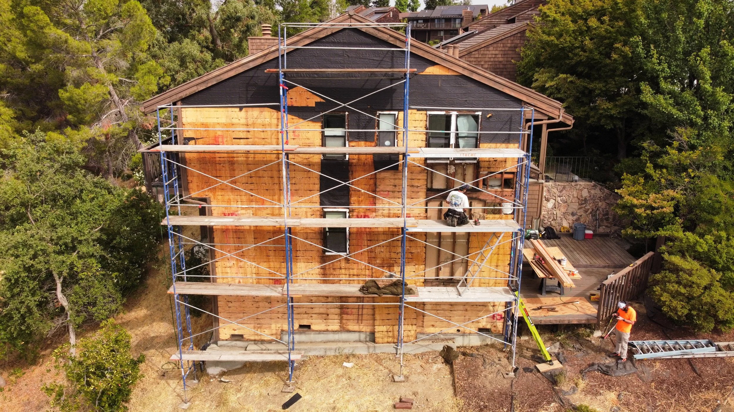A house under construction with scaffolding surrounding it. The house has part of its exterior wall covered with black weatherproof material, and other parts are exposed wood. Two workers are on the scaffolding, and one worker is on the ground near a ladder, working on the house.