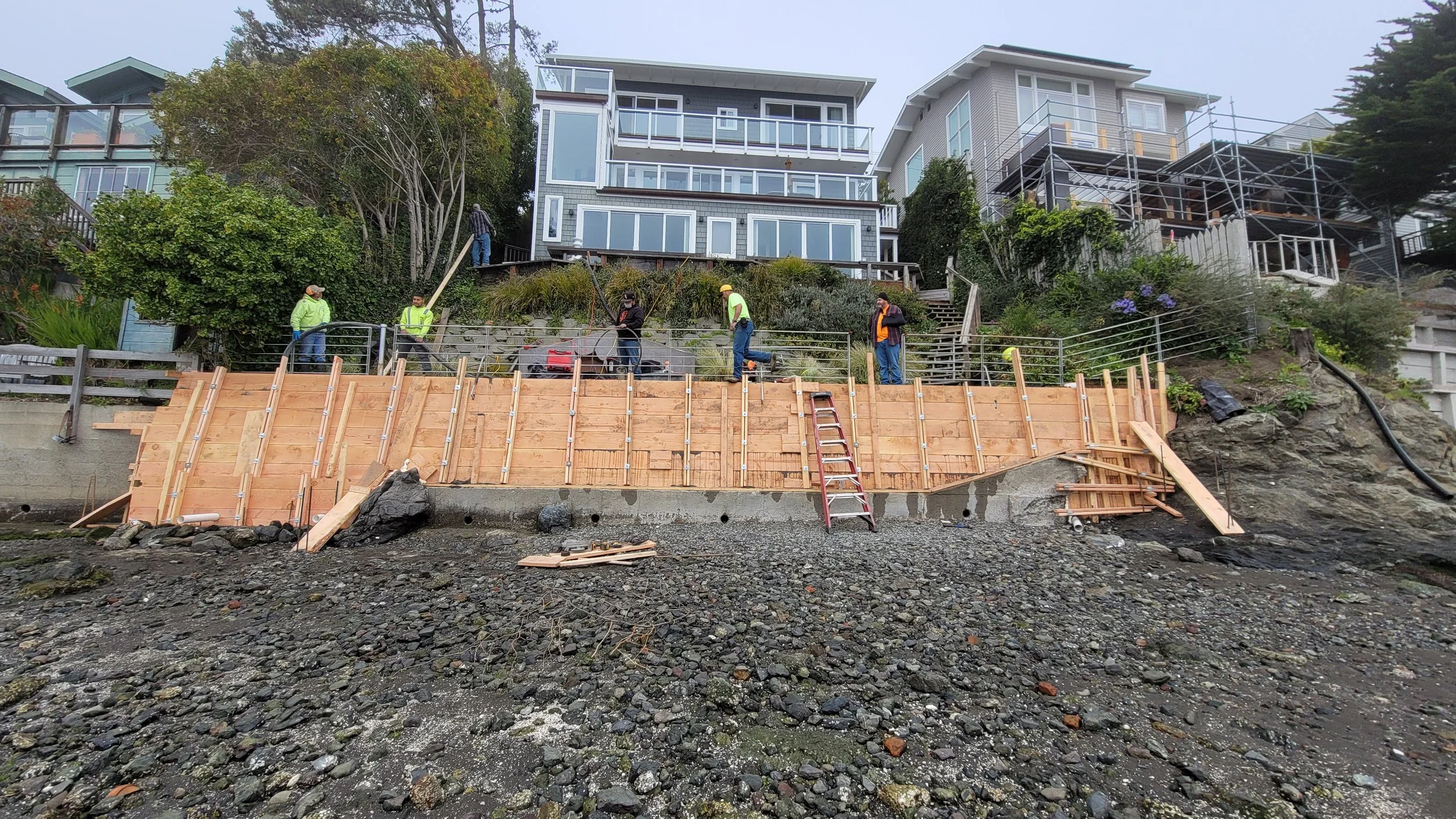 Construction workers building a wooden retaining wall on a rocky shoreline in front of modern houses on a hillside.