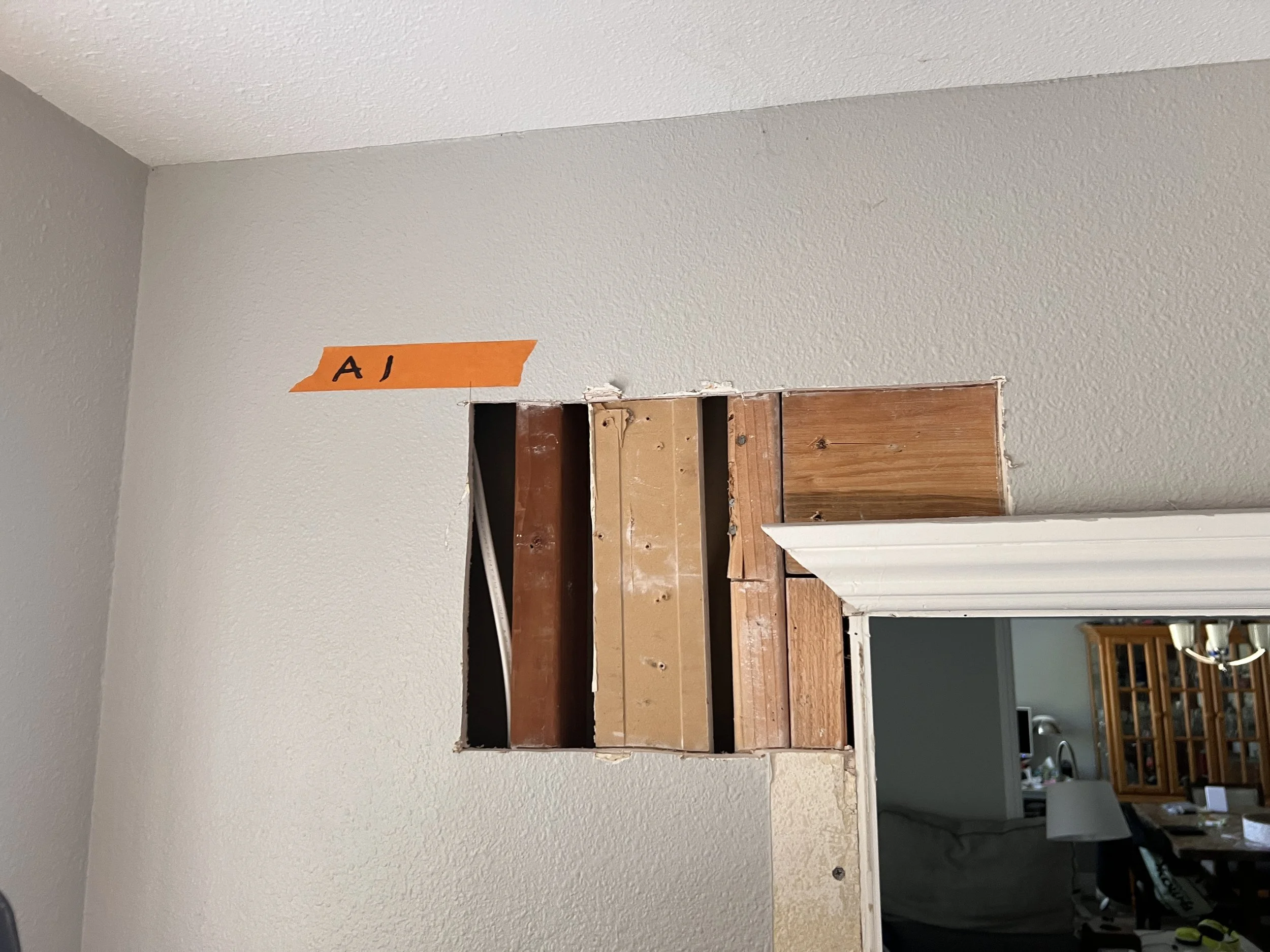 Interior wall with a cutout where drywall is removed, exposing wooden studs, with an orange tape labeled 'A J' on the wall.