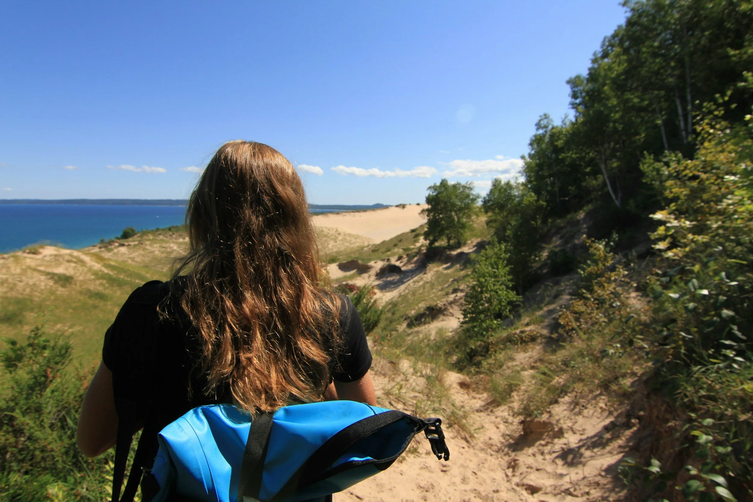 A person with long brown hair wearing a black shirt and a blue backpack walking on a dirt trail through a forested area near a body of water, under a clear blue sky with some clouds.