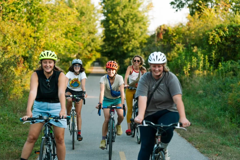 Group of five women biking on a paved trail surrounded by green trees and grass on a sunny day.