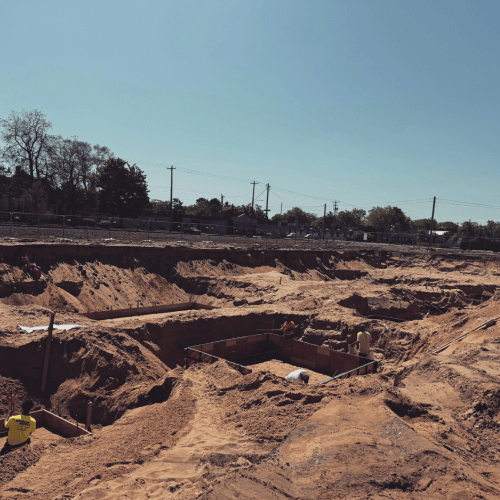 Construction site with a large excavation and concrete foundation, surrounded by dirt mounds, under a clear blue sky.