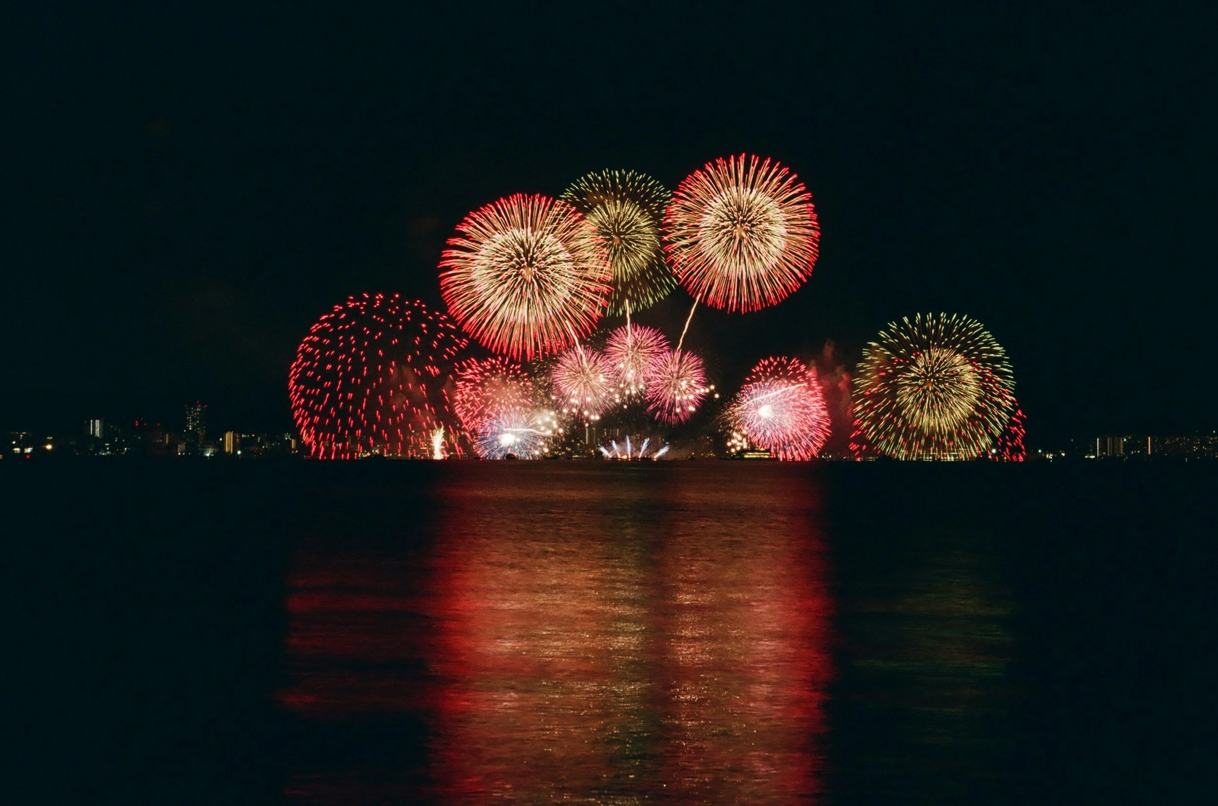Colorful fireworks display over a city skyline at night, reflected on water.