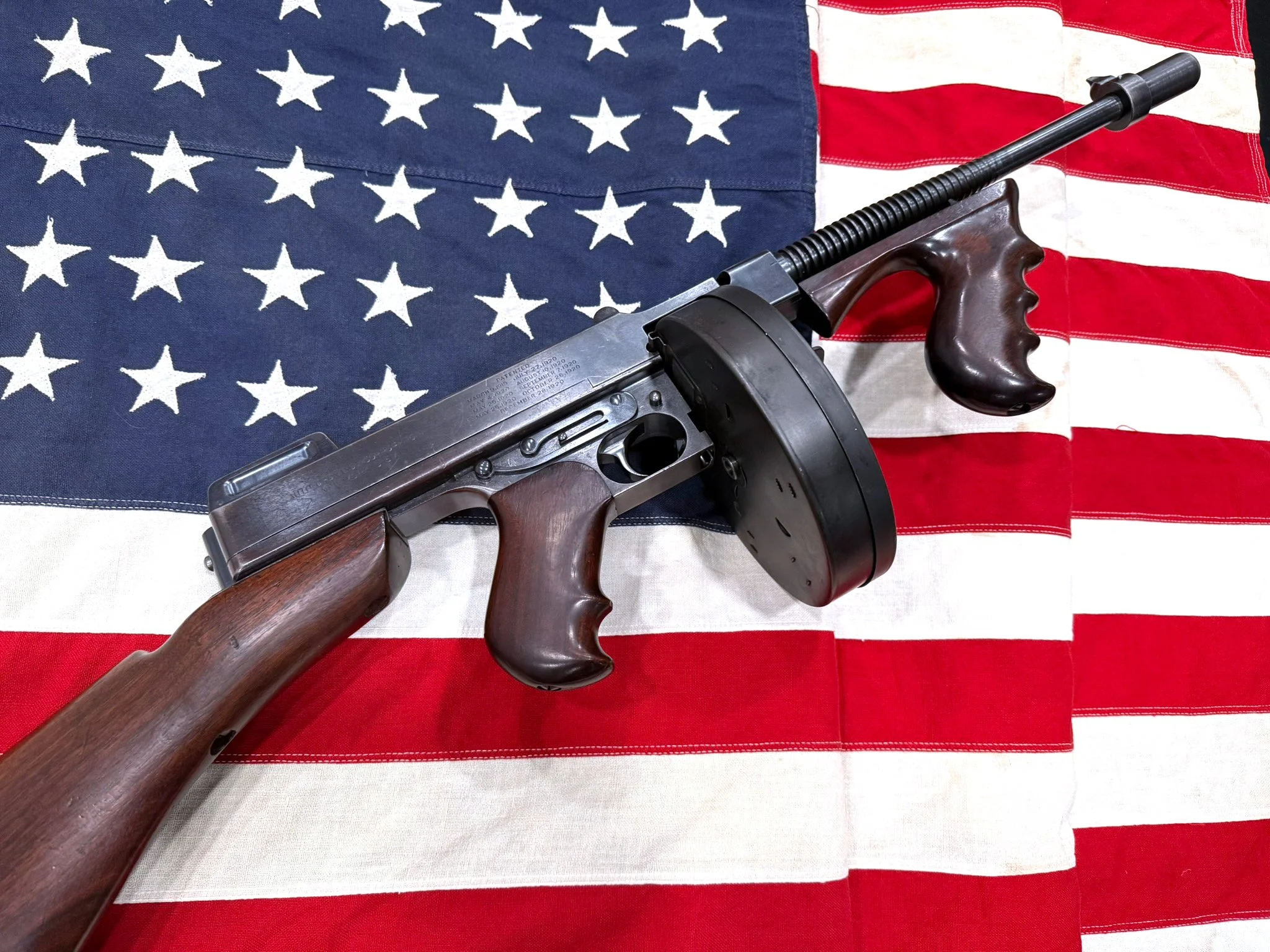 A vintage Thompson submachine gun with a drum magazine resting on an American flag backdrop.