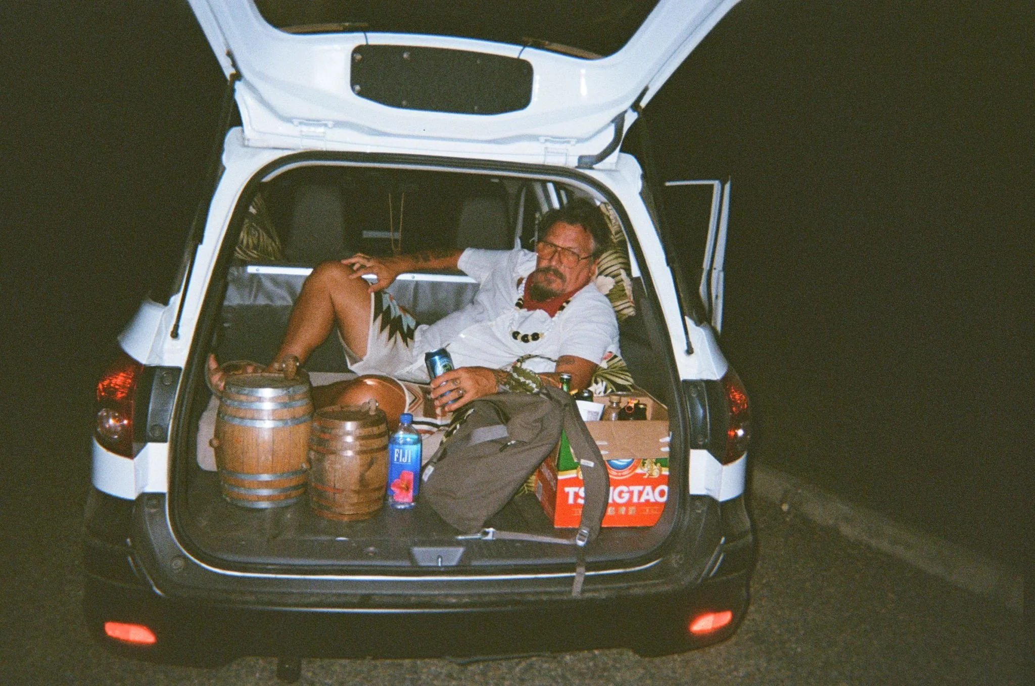 A man lying in the back of a parked car at night, surrounded by bottles and boxes, holding a drink and relaxing.