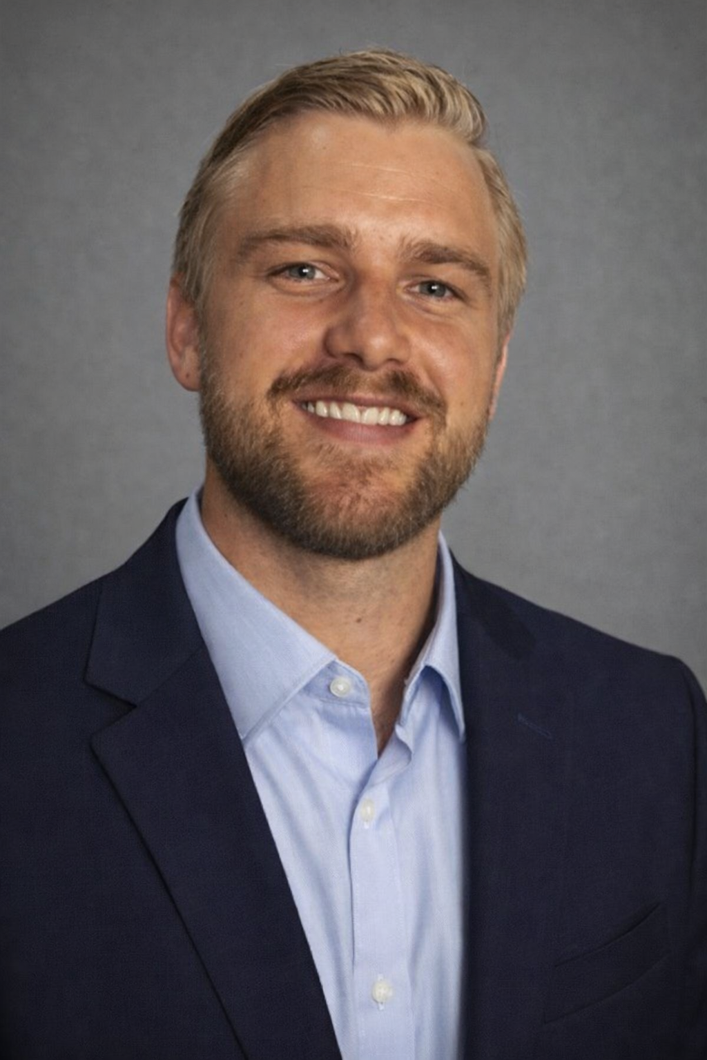 A professional headshot of a man with blonde hair and a beard, wearing a dark blazer and light blue shirt, smiling against a gray background.