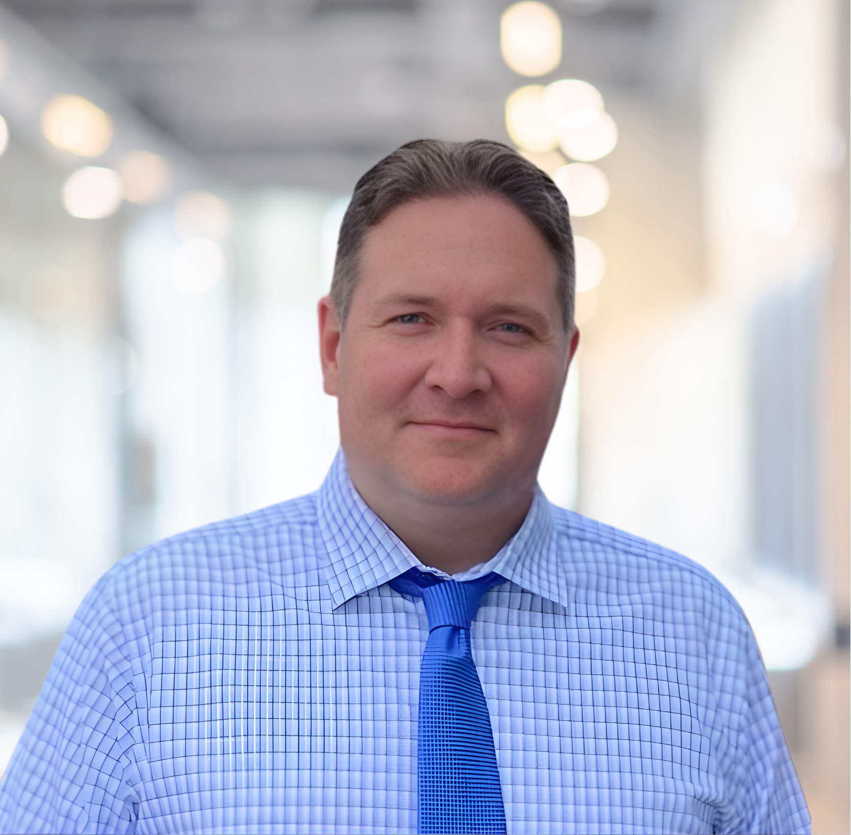 A man with short brown hair wearing a blue checkered shirt and a matching blue tie, standing in a brightly lit indoor space with blurred lights in the background.