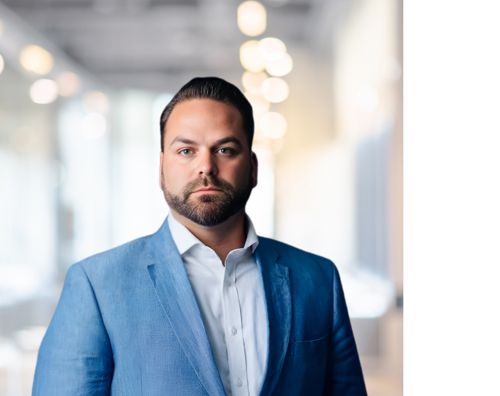 A man with dark hair, a beard, and light skin, wearing a blue suit and white shirt, standing indoors with blurred lights and windows in the background.