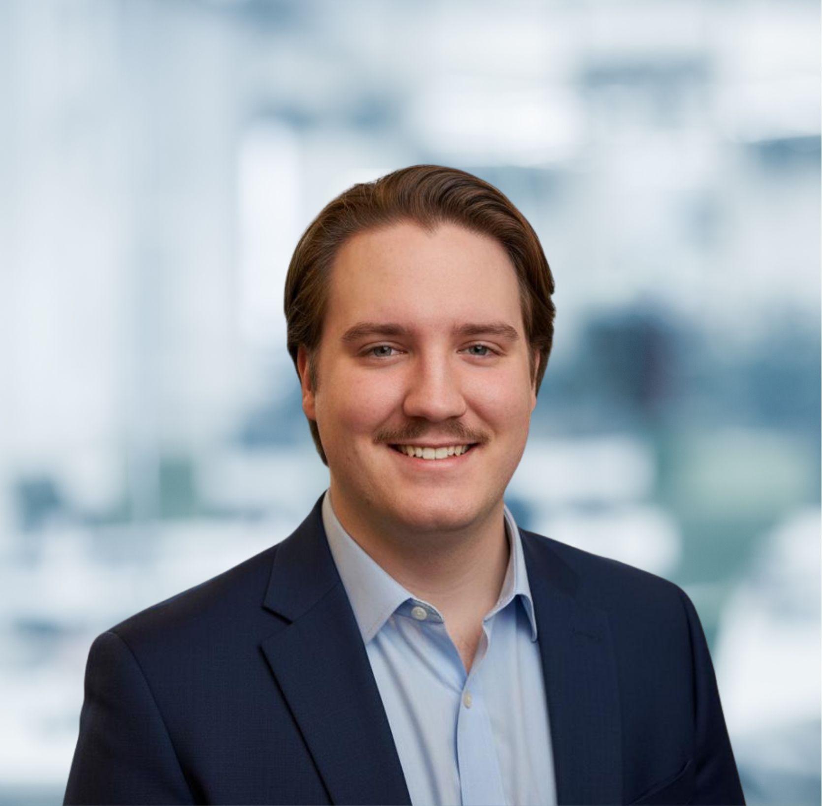 Portrait of a young man with brown hair, blue eyes, and a mustache, wearing a dark suit and light blue shirt, smiling in an indoor setting with a blurred modern office background.