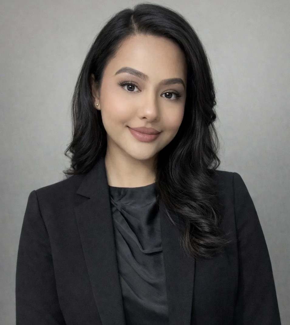Portrait of a young woman with dark, wavy hair smiling at the camera in an office setting with computers and a potted plant.