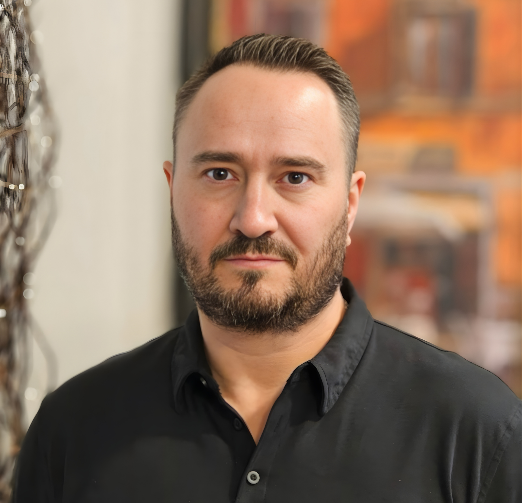 Close-up portrait of a man with short dark hair, beard, and mustache, wearing a black collared shirt, standing indoors with blurred artwork in the background.