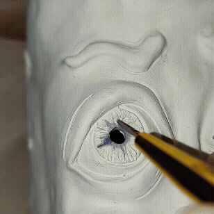 video description: artist carefully painting vibrant speedball glaze onto the detailed, textured surface of a ceramic sculptures eye