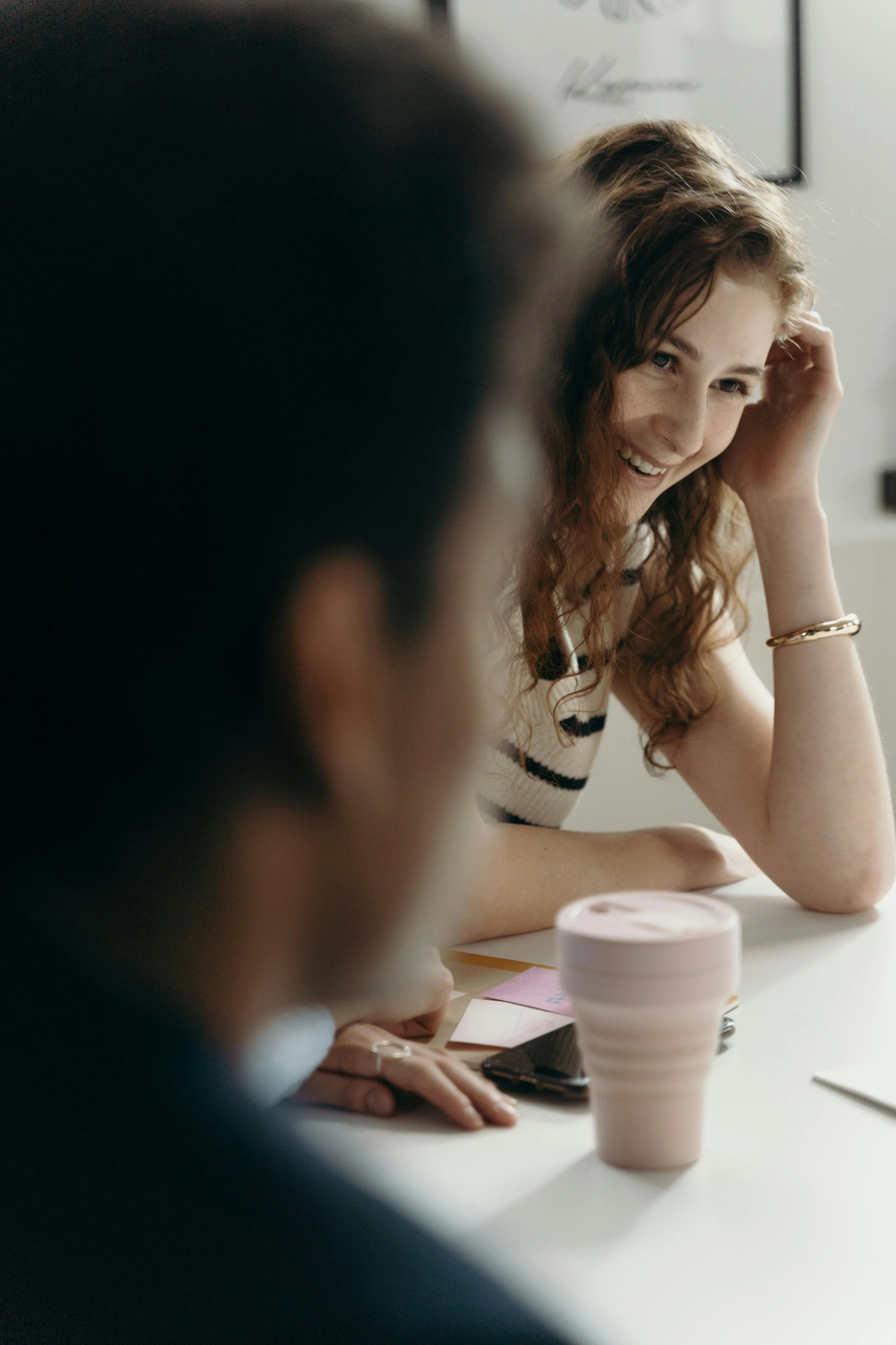 Mujer de cabello rizado y alegre en una mesa, conversando con otra persona cuyo rostro no es visible, con una taza rosada y un teléfono en la mesa.
