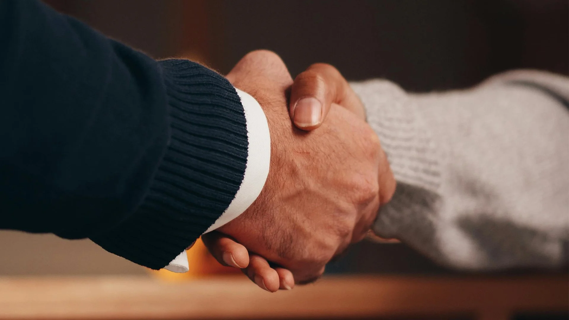 A close-up of two people shaking hands, one wearing a dark sweater and the other in a light gray sleeve.