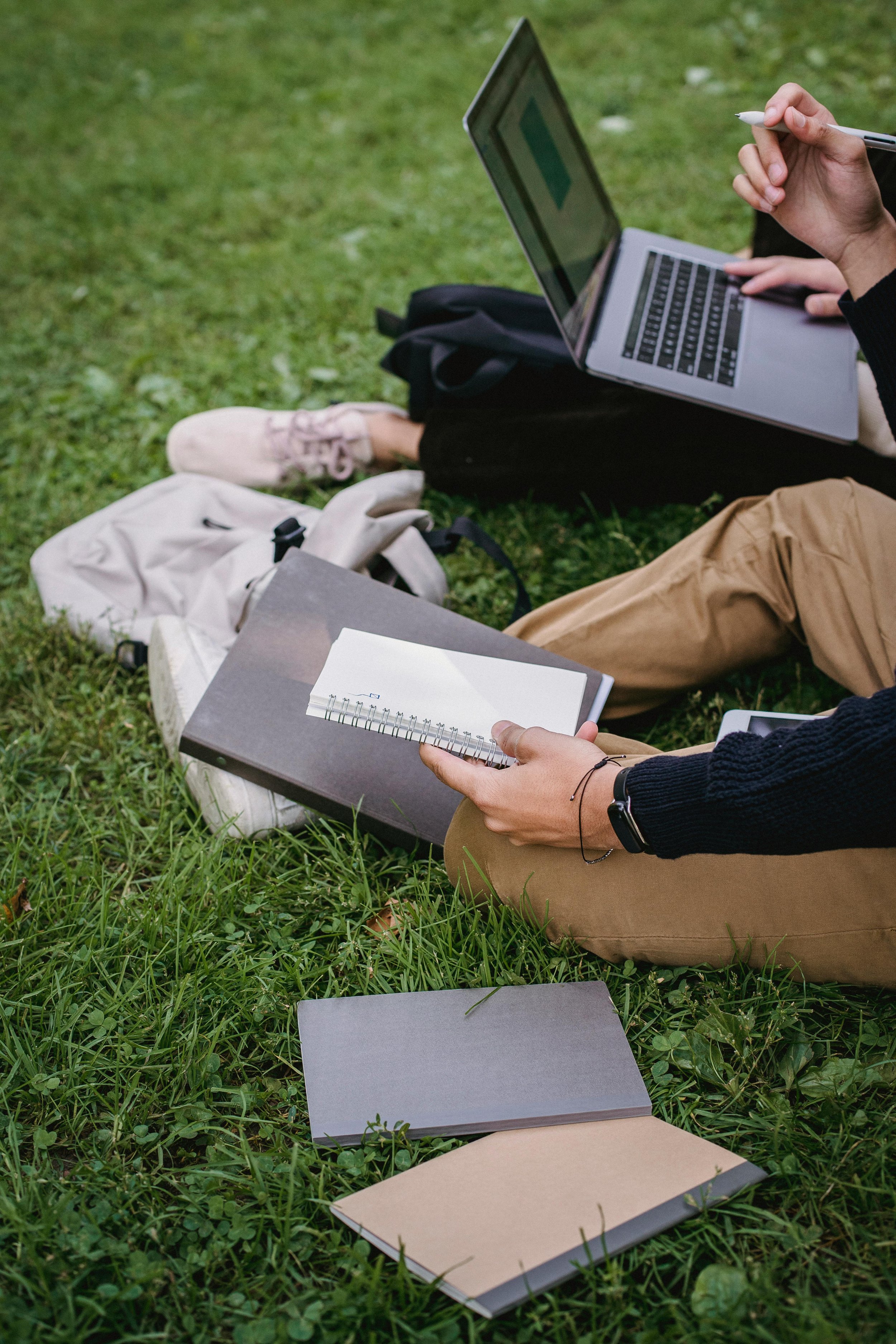 Personas sentadas en la hierba con laptops, cuadernos y una mochila, en un entorno al aire libre.