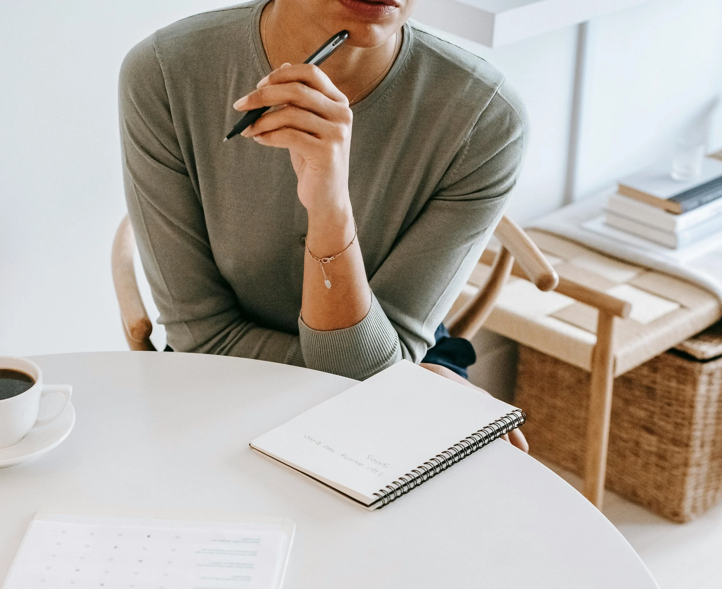 Mujer pensativa con bolígrafo en mano, sentada en una mesa blanca con una taza de café y libreta, en un ambiente de oficina o estudio, con estantes y libros en el fondo.