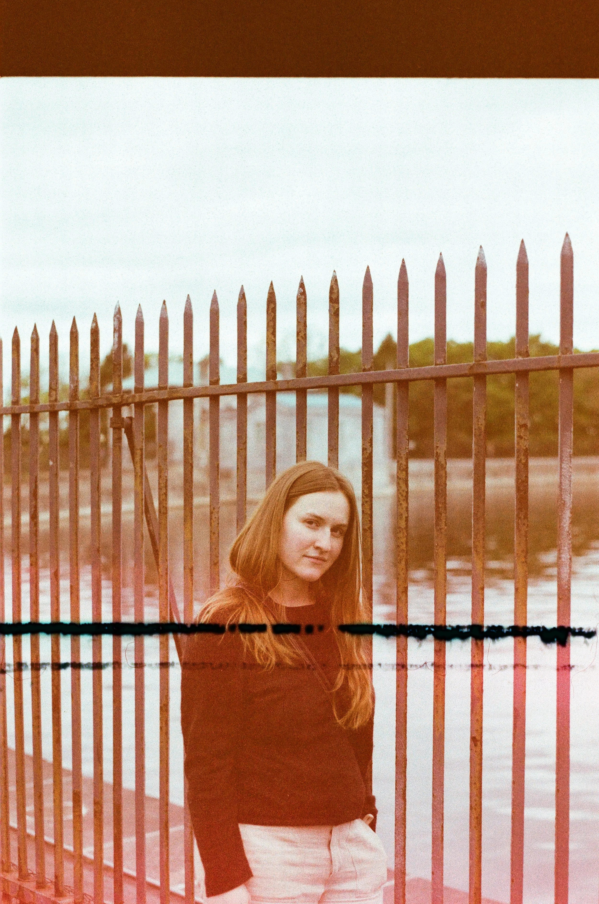 A young woman with long brown hair standing near a rusted metal fence beside a body of water, with trees in the background during daytime.