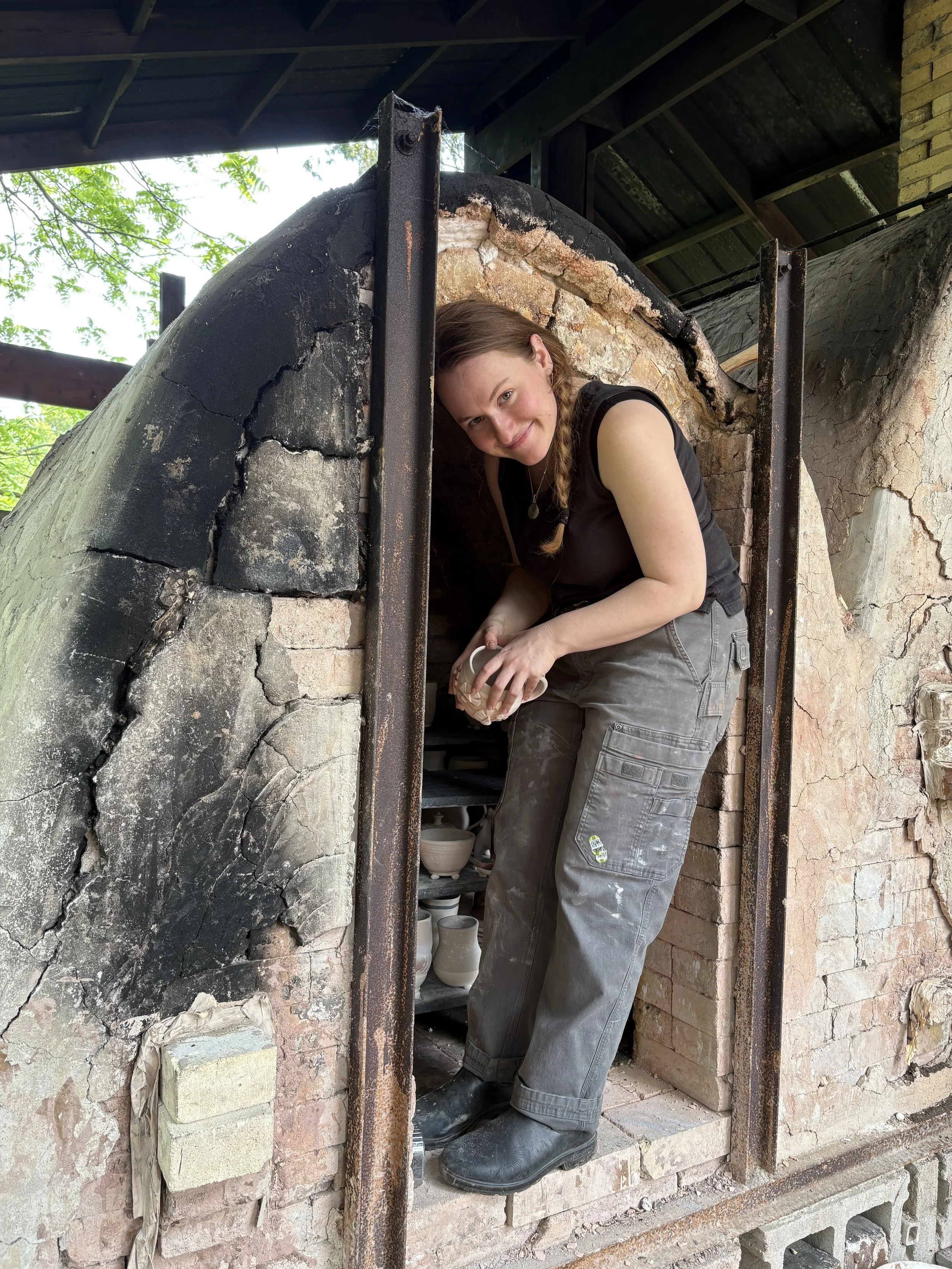 A woman with long braided hair, wearing a black sleeveless top, gray work pants, and black boots, is crouching inside a small brick and stone kiln or oven, holding a pottery piece she has just crafted.