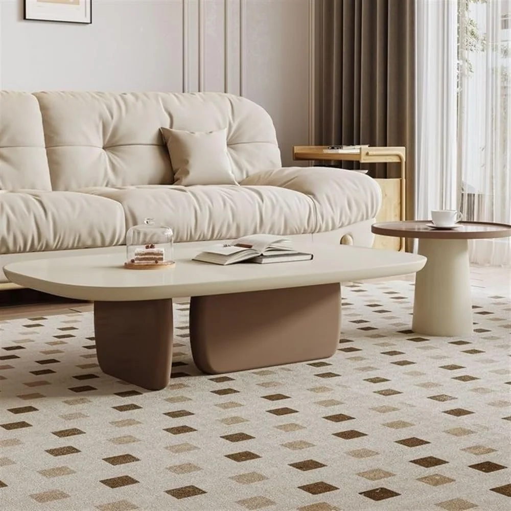 A living room with a cream-colored tufted sofa, a rectangular coffee table with books and a cake under a glass dome, a round side table with a mug, beige curtains, and natural light coming through sheer curtains.