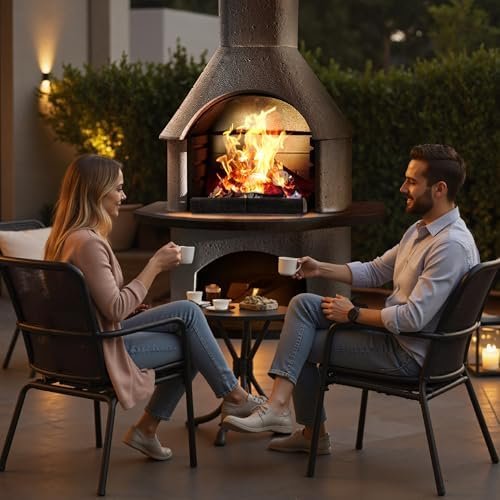 A man and woman sitting outside at a table, smiling and holding cups, with a large outdoor fireplace with a roaring fire behind them, surrounded by greenery and warm lighting.