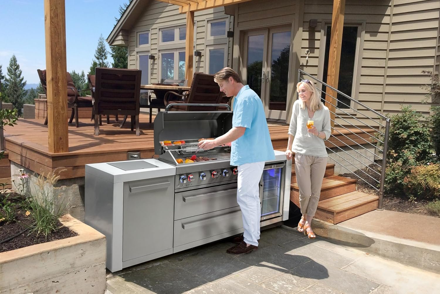 A man cooking food on an outdoor grill while a woman stands nearby holding a drink, on a patio outside a house with a large deck and outdoor furniture.