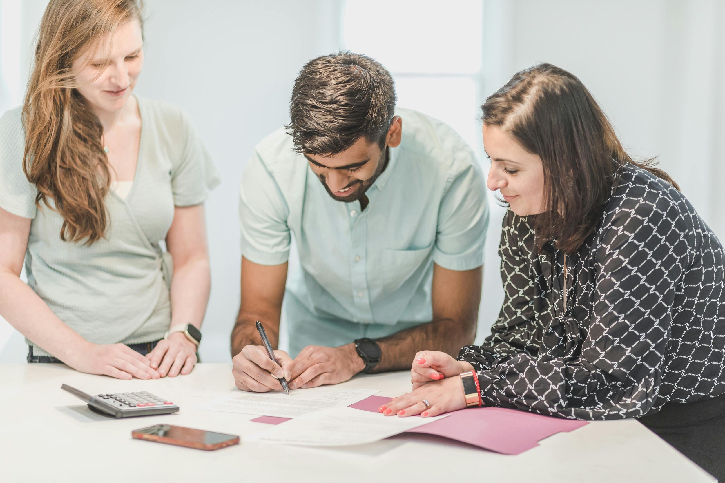 Three people are gathered around a table, engaged in a discussion or signing documents, with office supplies like a calculator, papers, and a phone nearby.