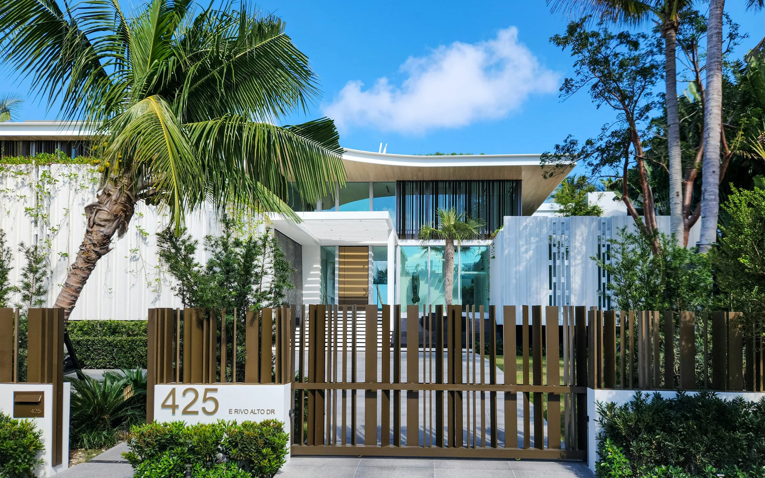 Modern house with a white fence, gate, palm trees, and lush greenery, situated at 425 E Rivo Alto Drive under a blue sky with clouds.