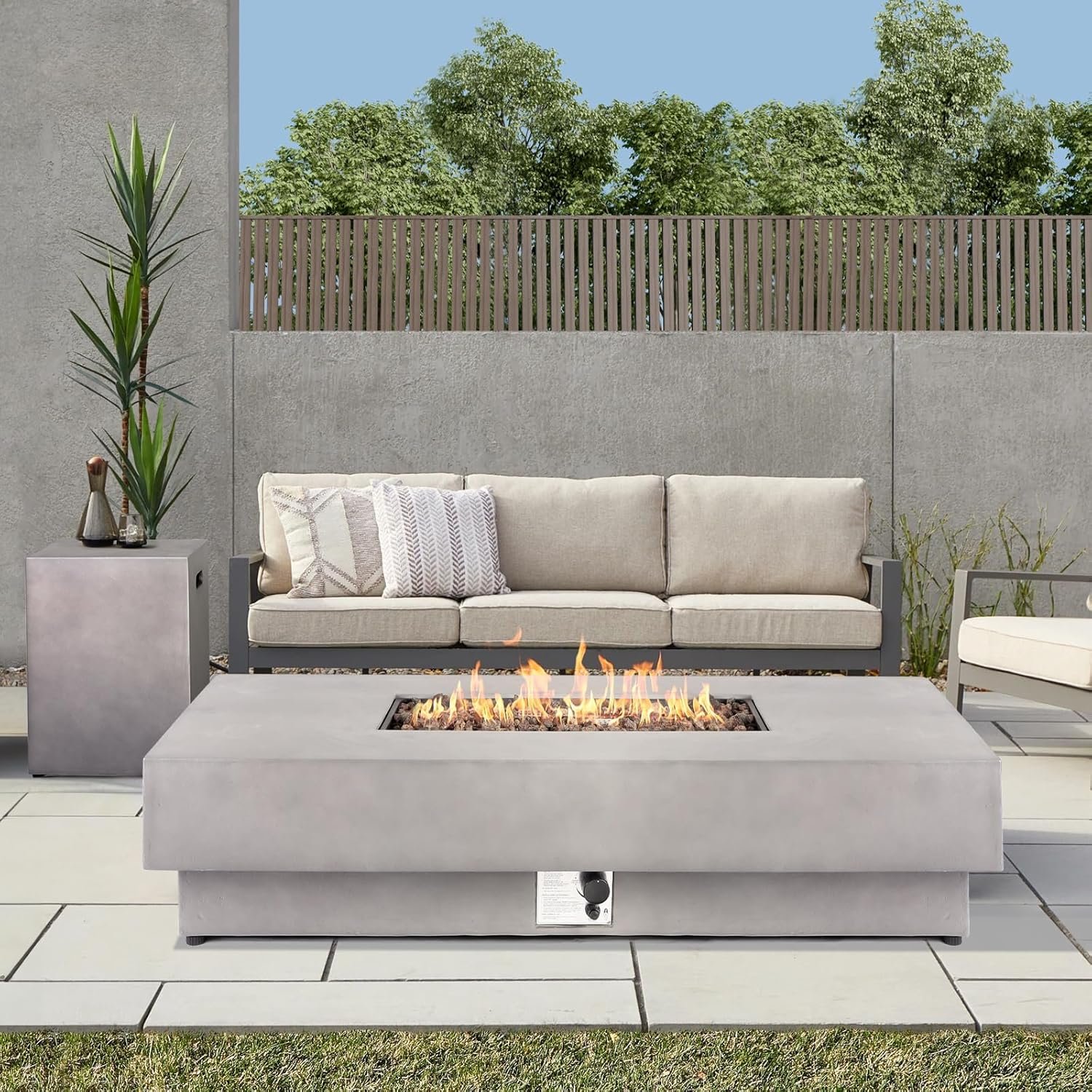 Outdoor patio with a concrete fire pit, a beige sofa with decorative pillows, and a side table with plants against a concrete wall and wooden fence, with green trees and blue sky in the background.